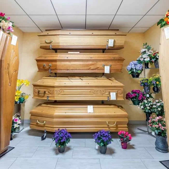 Quatre cercueils en bois clair exposés sur des rayonnages avec fleurs artificielles.